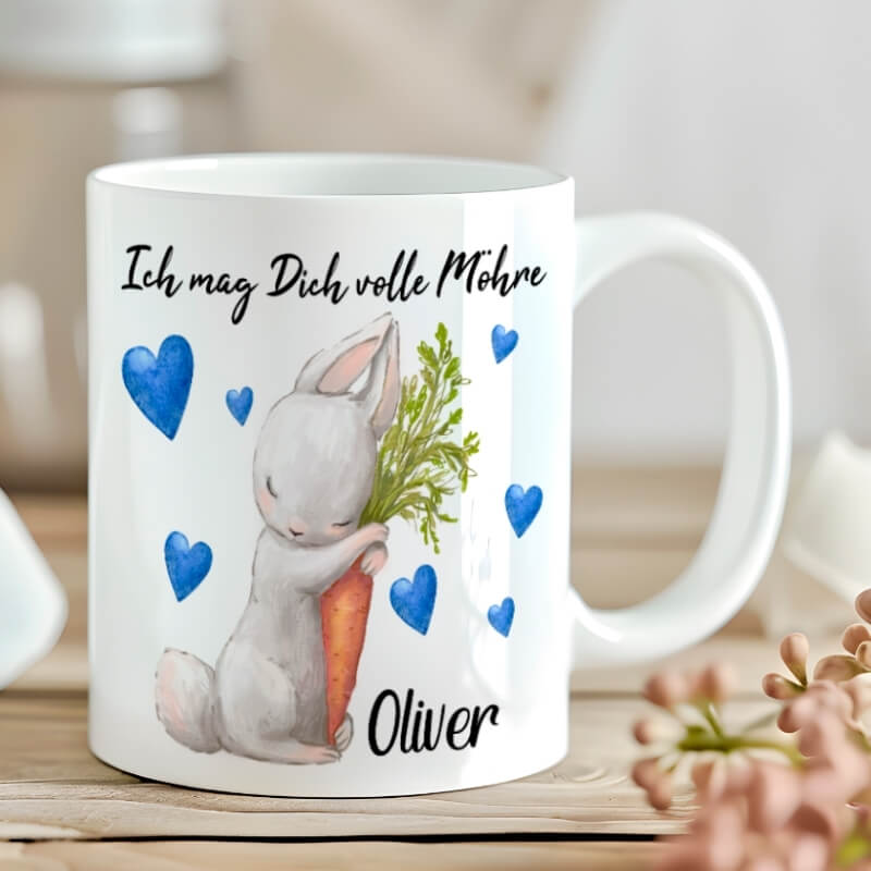 Tasse mit der Aufschrift „Ich mag Dich volle Möhre“ und blauem Herzdesign – personalisiert mit dem Namen Oliver, dekoriert mit Blumen im Hintergrund.