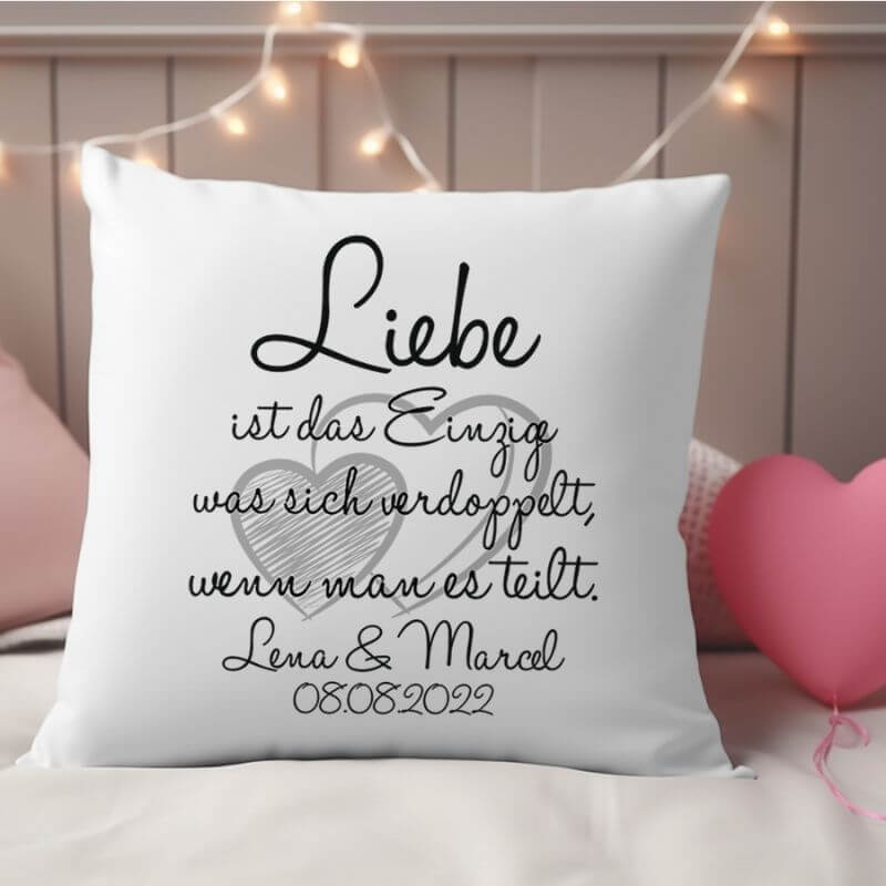 Weißes Kuschelkissen mit dem Aufdruck „Liebe ist das Einzige, was sich verdoppelt, wenn man es teilt“ und personalisierten Namen Lena & Marcel, liebevoll auf einem Bett mit Lichterkette gelegt.