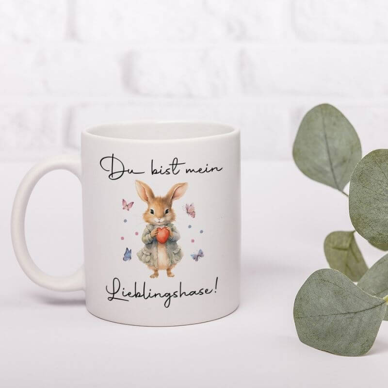 Tasse "Du bist mein Lieblingshase" steht auf einem weißen Tisch, rechts von der Keramiktasse eine grüne Pflanze.