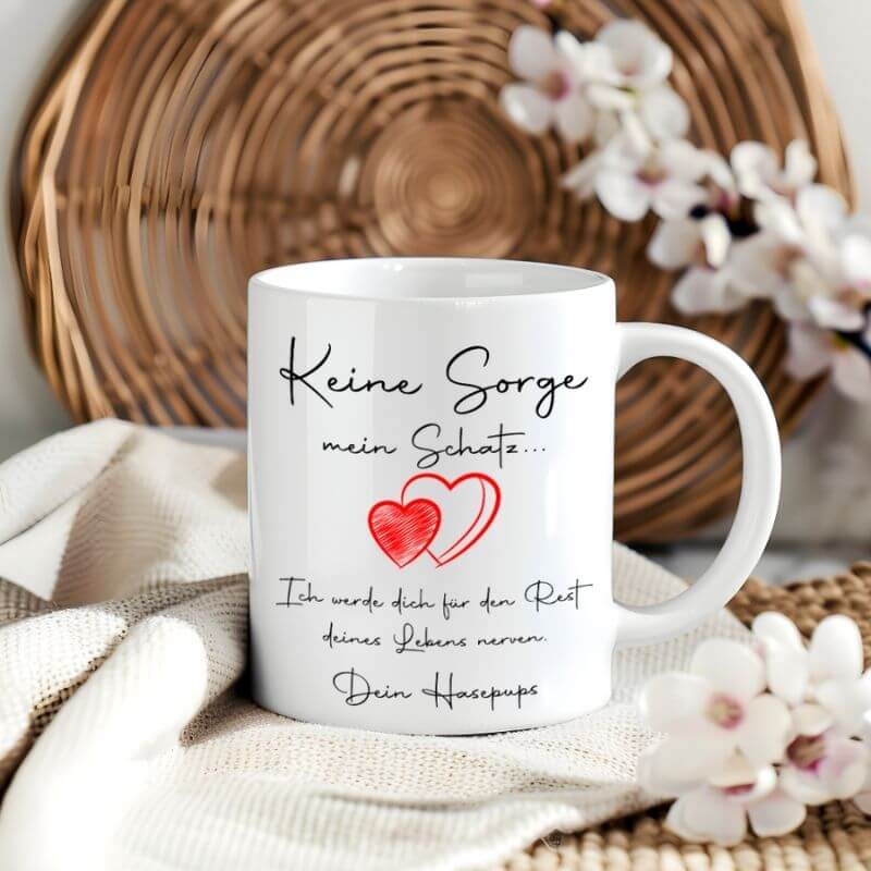 Weiße Liebestasse mit humorvollem Spruch "Keine Sorge mein Schatz, ich werde dich für den Rest deines Lebens nerven", aufgestellt vor einem Flechtkorb mit Blumen.