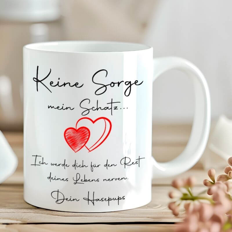 Personalisierte Tasse mit dem Spruch "Keine Sorge mein Schatz" und roten Herzen, liebevoll platziert auf einem Holztisch mit zarten Blüten im Hintergrund.
