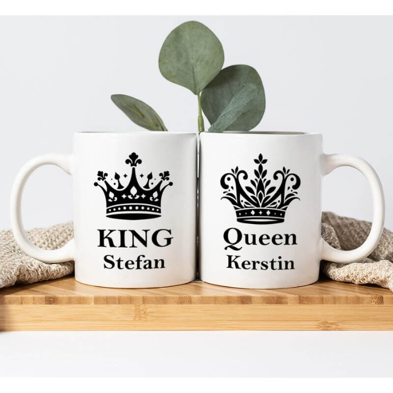 Ein weiteres Produktbild von den Partnertassen "King und Queen". Platziert auf einem Holztisch mit einer beigen Tischdecke und einer grünen Pflanze hinter den Tassen.