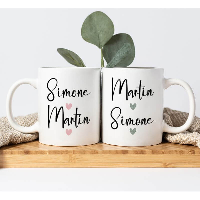 Beide Tassen stehen auf einem Holztisch. Hinter den Tassen liegt eine beige Tischdecke. Eine grüne Pflanze steht im Hintergrund von den Tassen.